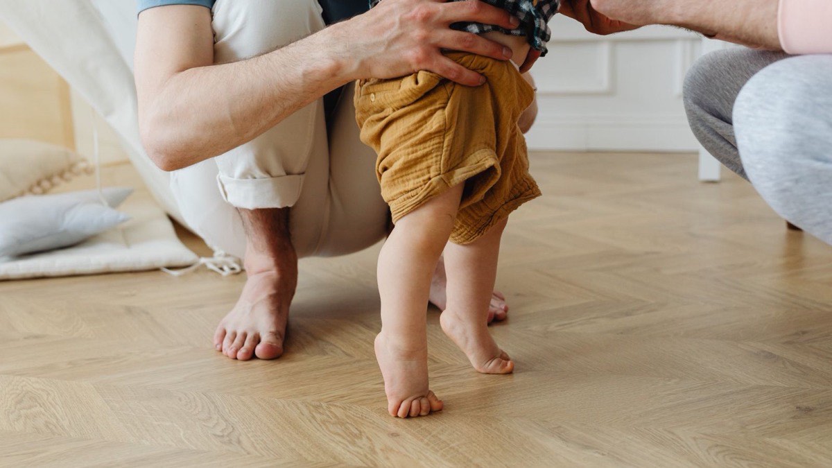 A baby taking first steps, held by a parent’s hands
